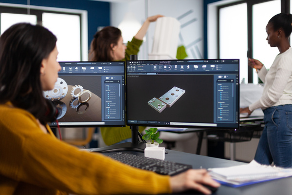 Industrial engineer woman working at pc with two monitors, screens showing CAD software with 3D architecture prototype of gears mechanical piece. Entrepreneur working in creative construction office